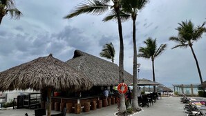 Un restaurante frente a la playa con techos de paja, mesas al aire libre y palmeras.