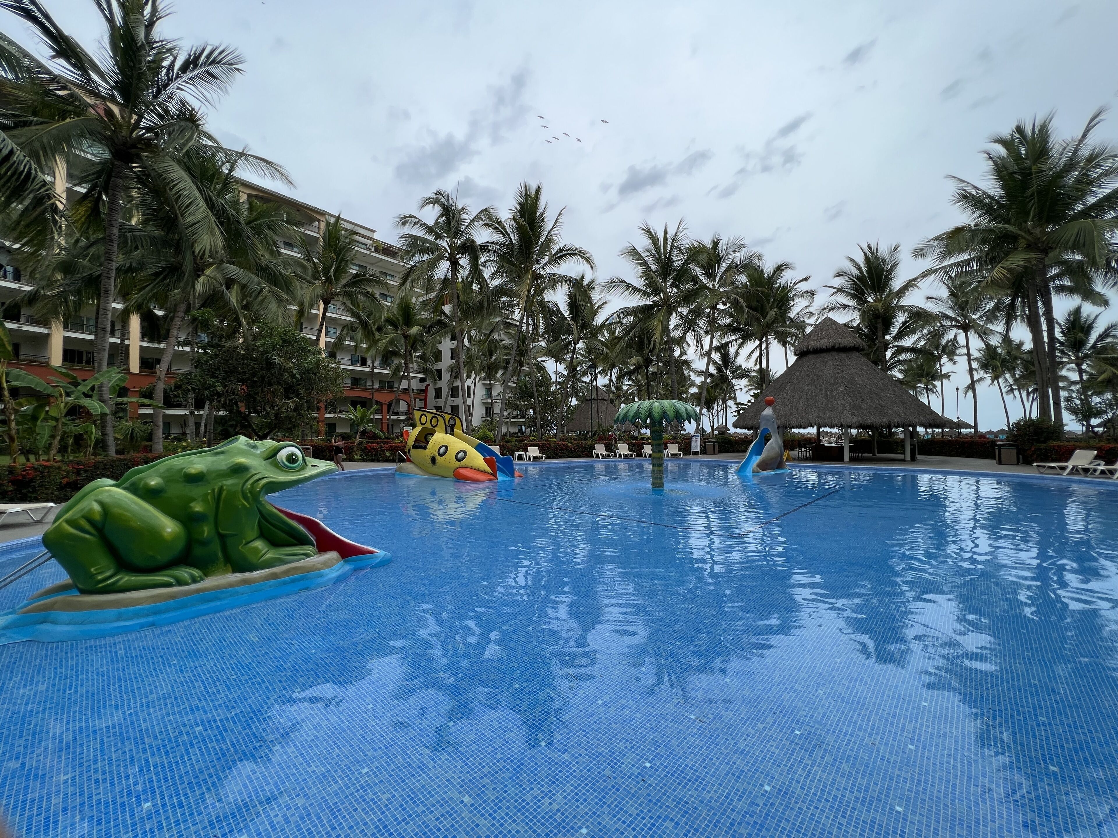 A swimming pool with a frog float, palm trees, and a thatched-roof hut.