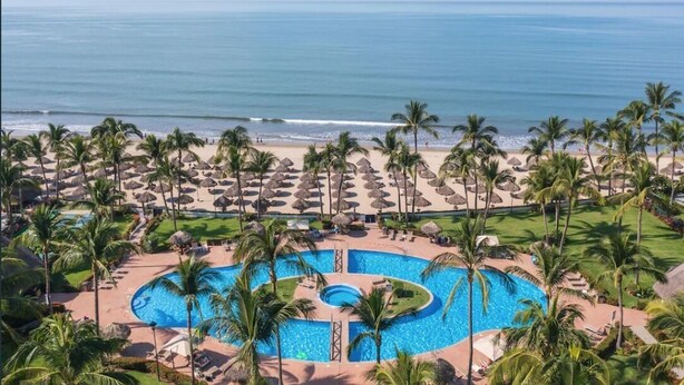 Un resort con piscina central, palmeras, sombrillas de playa y vistas al océano.