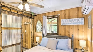 A wooden cabin bedroom with a bed, a nightstand, a window with curtains, a ceiling fan, and a sign that reads "NO WAKE NODFORK LAKE". - Remodeled Cabin w/ Fire Pit on Norfork Lake! (Elizabeth)