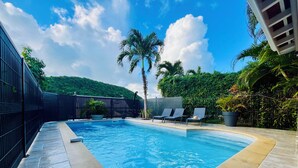 Ein rechteckiger Swimmingpool mit klarem, blauem Wasser, umgeben von einem schwarzen Zaun und einer gefliesten Liegefläche. Im Hintergrund sind Liegestühle und eine Palme zu sehen.