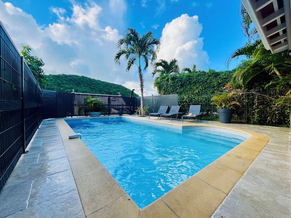 A rectangular swimming pool with clear blue water, surrounded by a black fence and a tiled deck. There are lounge chairs and a palm tree in the background. - Villa dO Sainte Anne, Piscine & Proche Plage (Sainte-Anne)