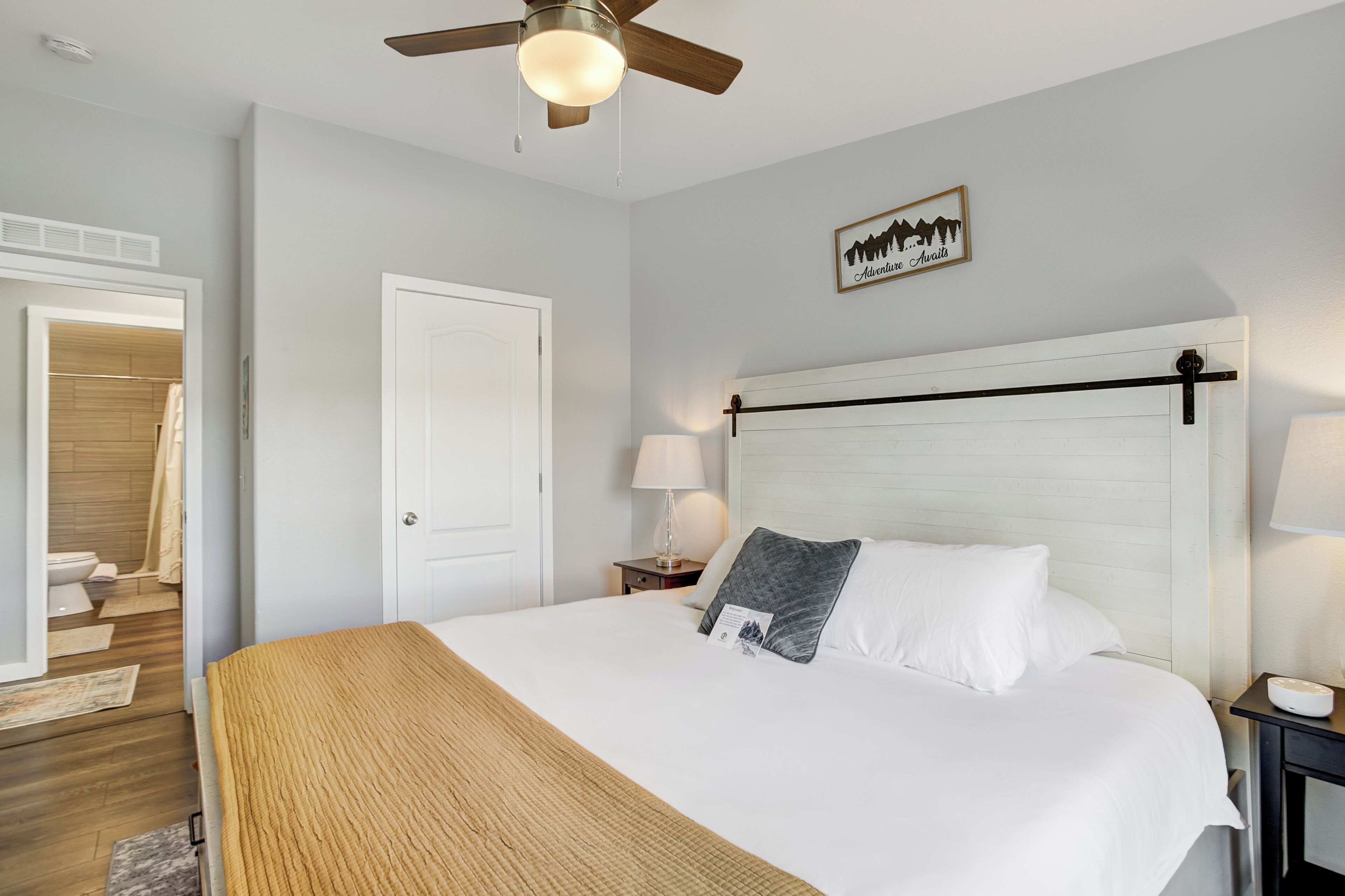 A hotel room with a bed, a ceiling fan, a nightstand with a lamp, a door, and a framed picture on the wall.