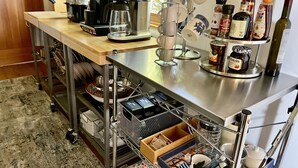 Una cocina con una cafetera, tazas de café, un tarro de mermelada y una botella de aceite de oliva sobre la encimera.