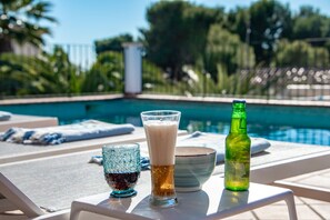 Een zwembadterras met ligbedden, een biertje, een glas donkere vloeistof en een bierfles op een tafel.