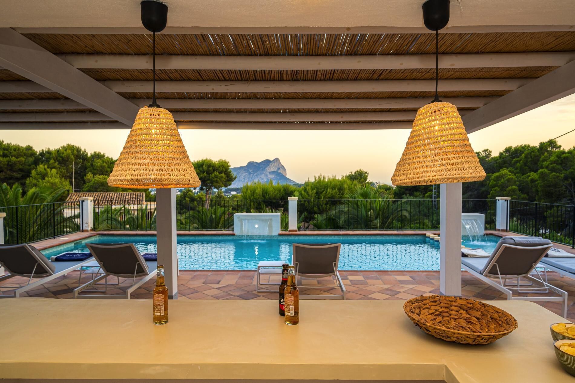 A poolside area with two wicker lamps, lounge chairs, and a table with bottles and a basket.