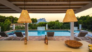 Área da piscina com duas luminárias de vime, espreguiçadeiras e uma mesa com garrafas e um cesto.