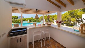 Cozinha com churrasqueira embutida, janela com vista para piscina e varanda com cadeiras e vista para o verde ao redor.