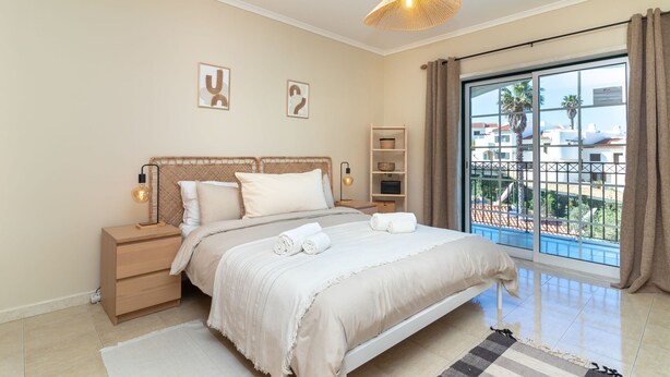 A bedroom with a bed, bedside tables, a lamp, a shelf, and a view of buildings through a window. - Casa Bela Vista - Communal pool, close to town centre, beach and amenties (Carvoeiro)