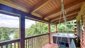 Una terraza de madera con un jacuzzi y una silla colgante con vistas a un área boscosa y un lago.