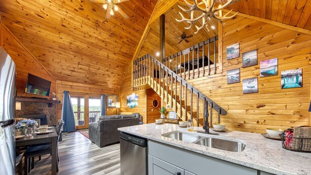 Interior de una cabaña con revestimiento de madera, una cocina con encimeras de granito, un comedor y una escalera que lleva a un altillo.