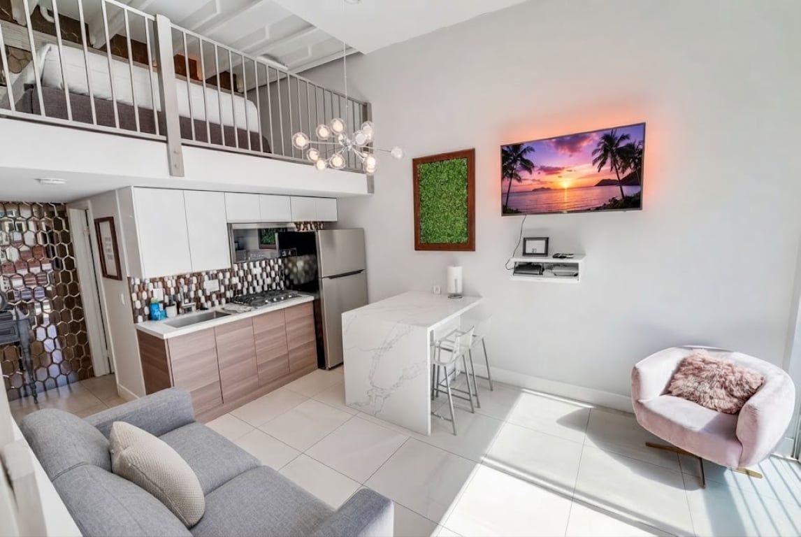 Une cuisine moderne avec un îlot en marbre, un coin repas avec une table et des chaises blanches, un salon avec un canapé et un balcon doté d’une balustrade.