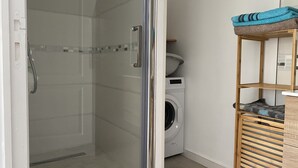 A bathroom with a glass shower enclosure, a washing machine, and a wooden shelf with towels. - Apartment in the heart of the wild south (Saint-Joseph)