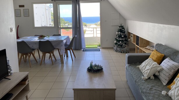 A spacious living area with a dining table, a sofa, and a view of the sea through a large window. - Apartment in the heart of the wild south (Saint-Joseph)