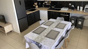 A compact kitchen with a refrigerator, stove, sink, and dishwasher. There is a dining table with chairs and a patterned tablecloth. - Apartment in the heart of the wild south (Saint-Joseph)