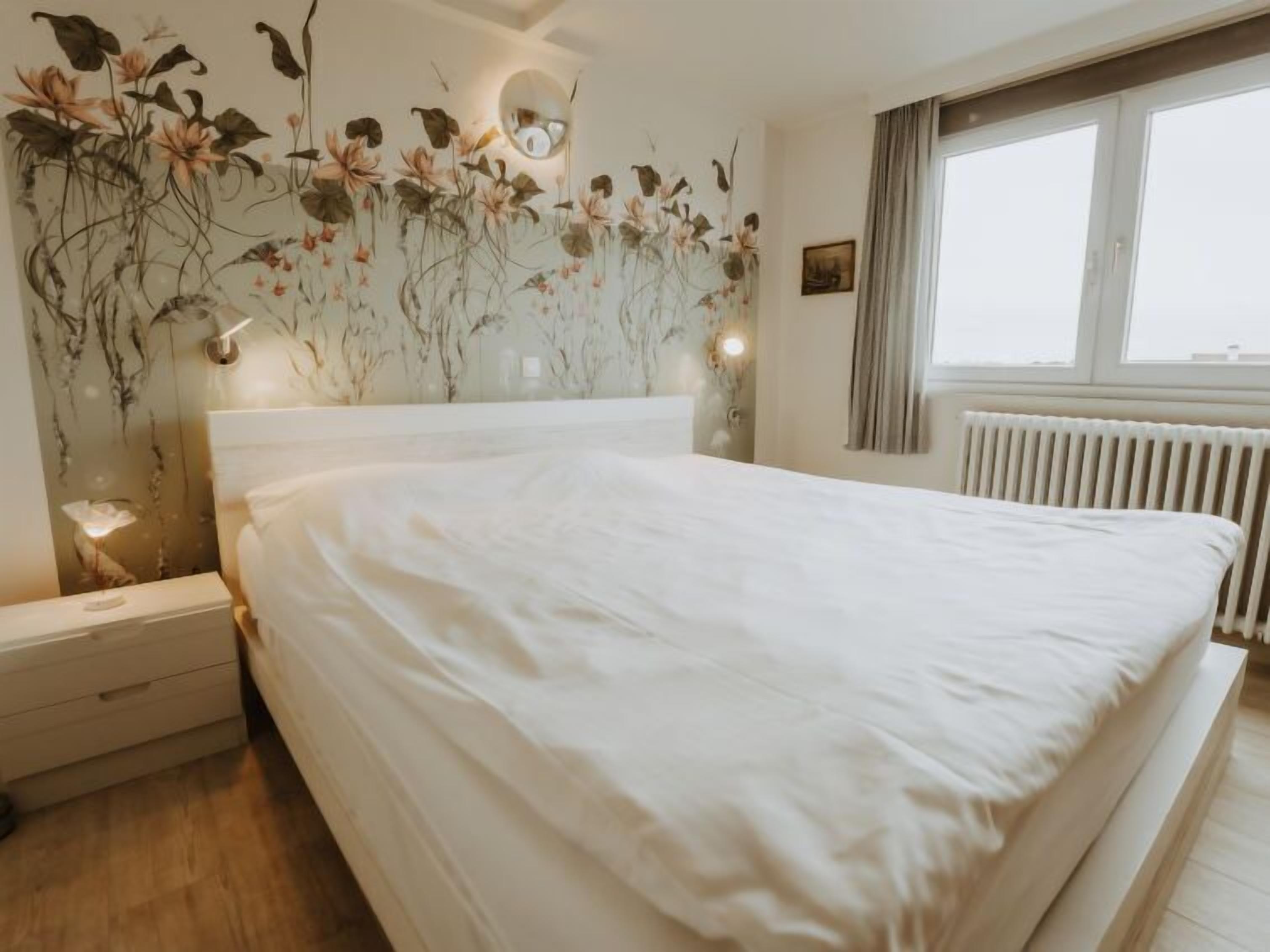 A bedroom with a large bed, a nightstand, a wall-mounted lamp, and a window with curtains.