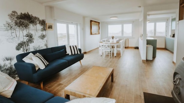 Un vaste espace de vie comprenant un canapé bleu foncé, une table basse en bois et une table à manger avec des chaises. On aperçoit également un coin cuisine avec une cuisinière et des placards.