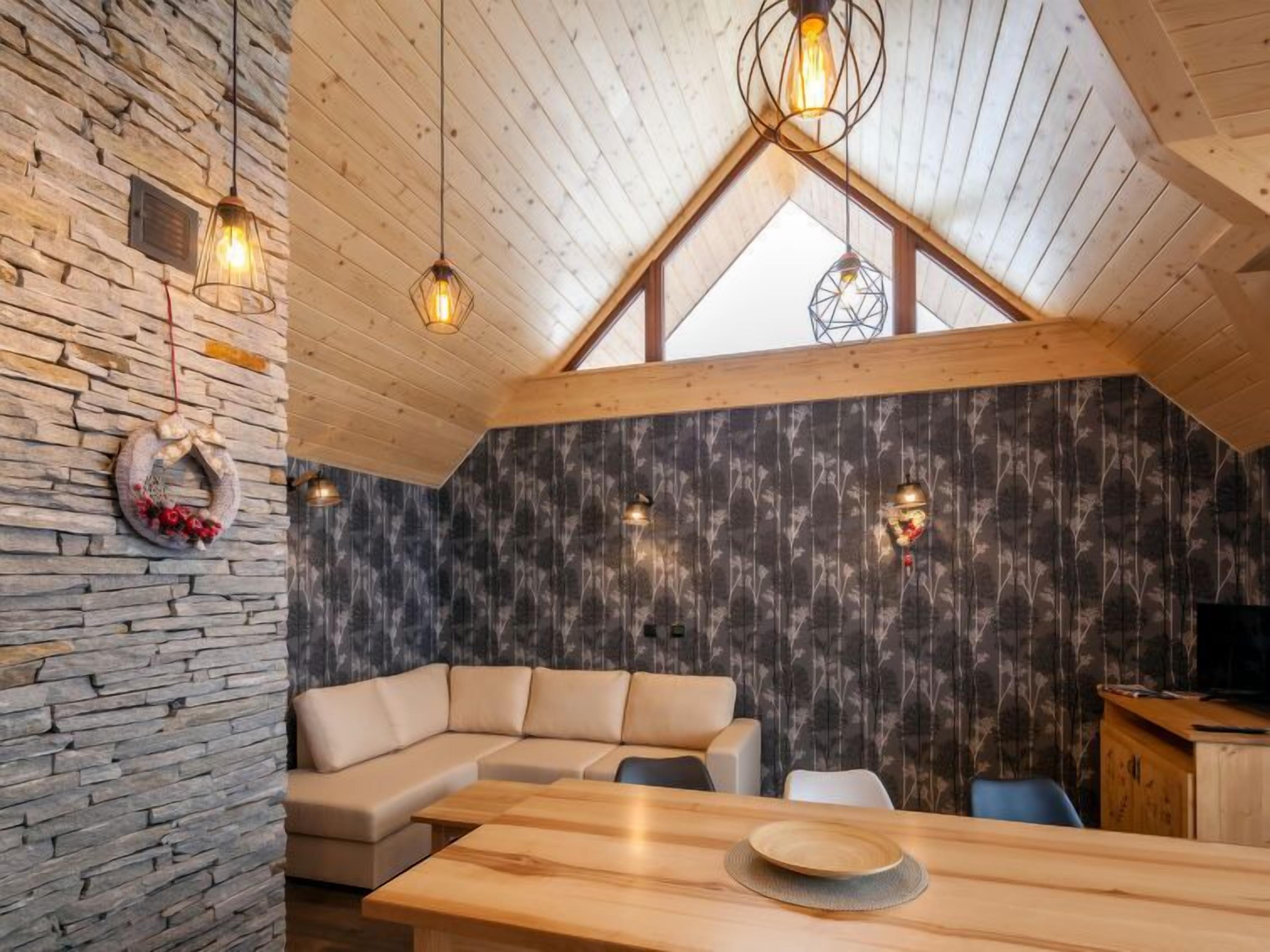 A cozy room with a stone wall, a wooden ceiling, and a dark patterned wall. There is a beige sofa, a wooden table, and a TV on a wooden cabinet.