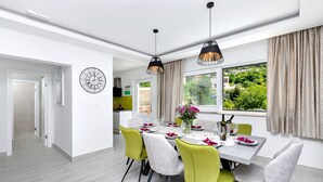 Un comedor con una mesa preparada para cuatro personas, rodeada de sillas con tapicería en tonos verde y gris. Hay lámparas colgantes sobre la mesa, y una ventana con cortinas a la derecha.