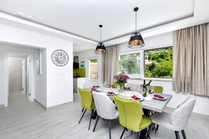 A dining area with a table set for four, surrounded by chairs with a mix of green and grey upholstery. There are pendant lights above the table, and a window with curtains on the right. - Villa Vivace with pool (Makarska)
