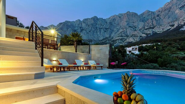 Zona de piscina con escalones que conducen a una terraza, sillas de descanso y una cesta de frutas.