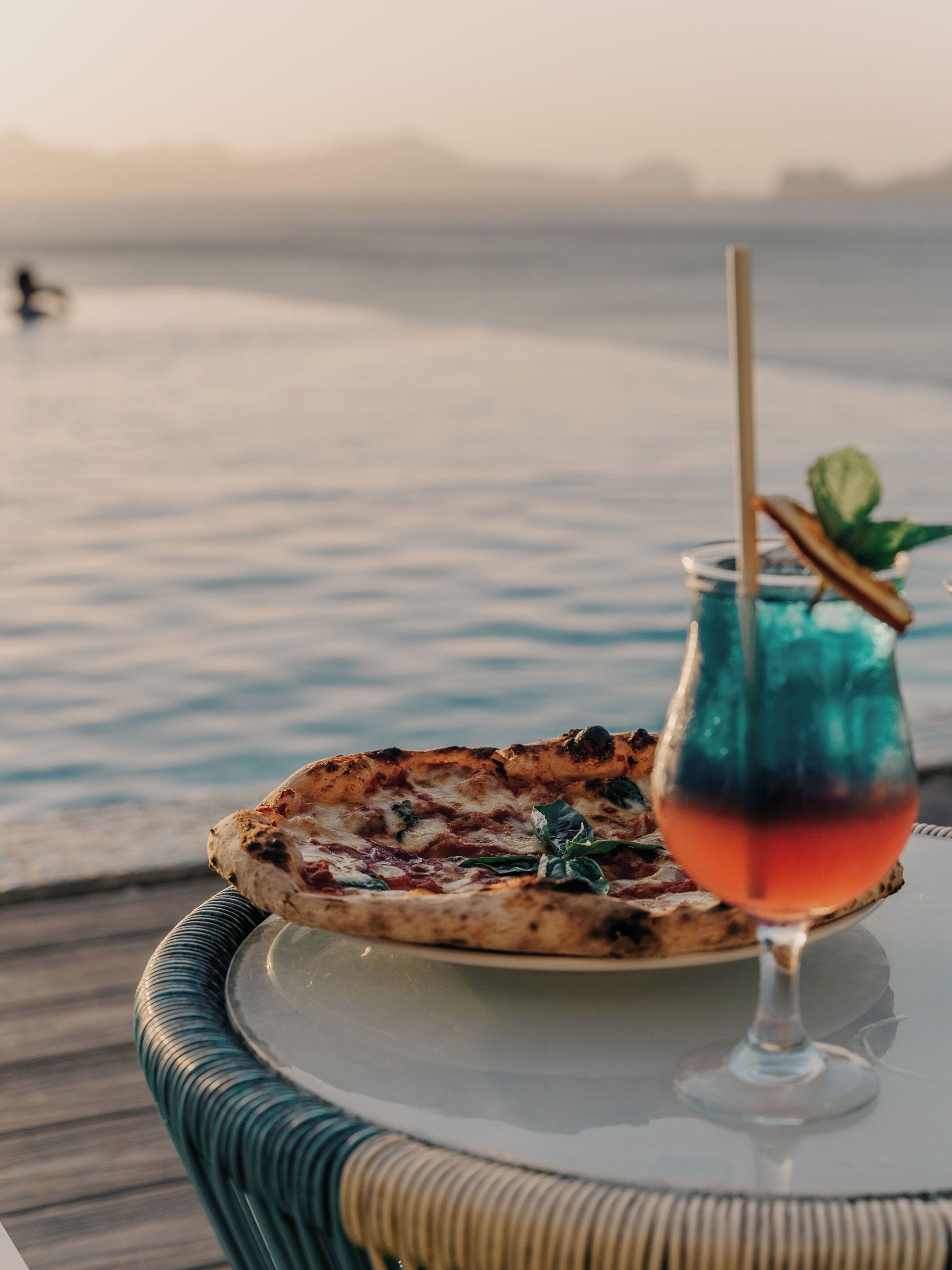A pizza on a table with a glass of drink and a view of water.