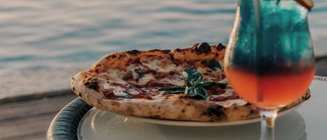 A pizza on a table with a glass of drink and a view of water.