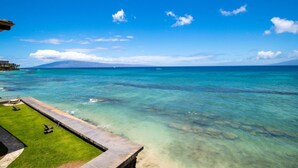 A clear view of a pool with a grassy area and lounge chairs, overlooking a turquoise sea with a distant mountain range.