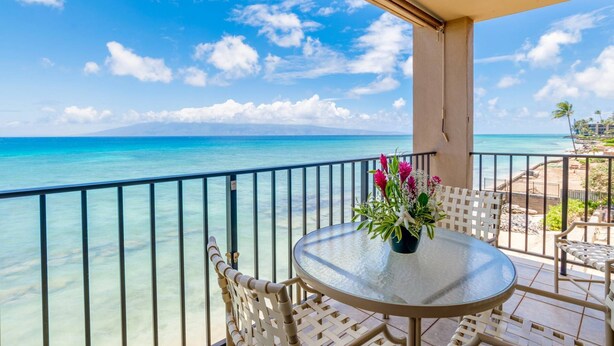 A balcony with a glass table and chairs, overlooking a beach with clear turquoise water and a distant mountain.