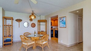 A dining area with a round table and four wicker chairs, a hanging light fixture, and a kitchen visible through an open doorway.