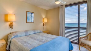 A hotel room with a bed, two wall-mounted lamps, a ceiling fan, a large window with blinds, and a view of the ocean.