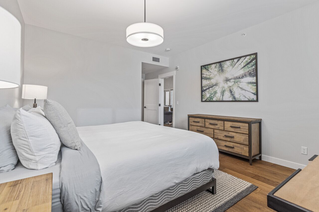 Een moderne slaapkamer met een groot bed, een houten dressoir, een abstract schilderij aan de muur en een hanglamp.
