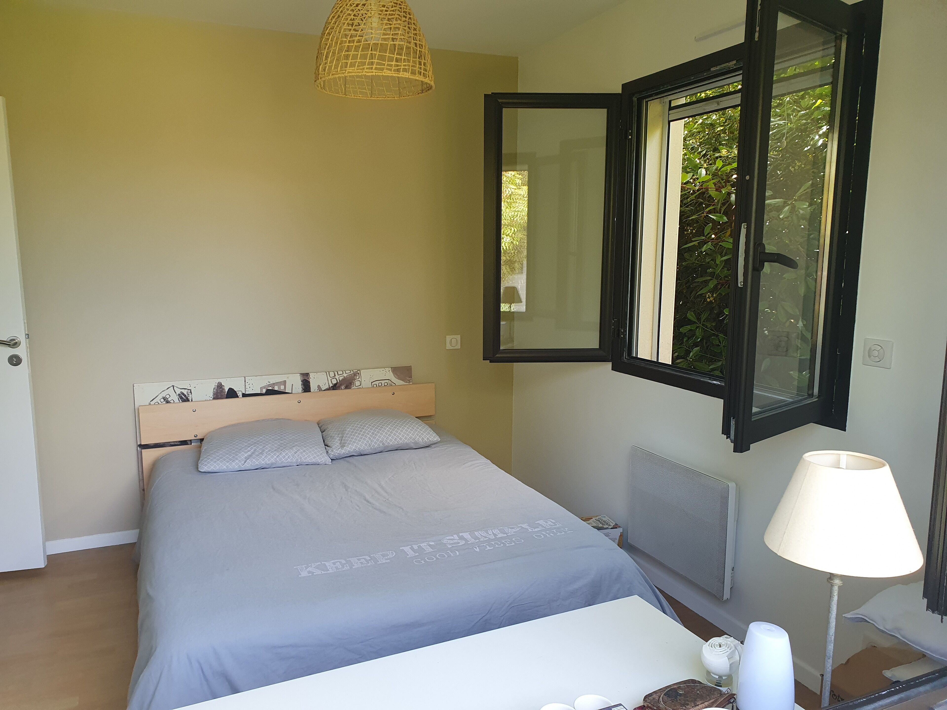 Une chambre à coucher comprenant un lit, une table de chevet, une lampe, une fenêtre avec un volet ouvert et un miroir fixé au mur.