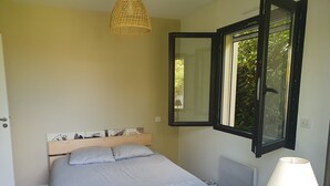 Un dormitorio con cama, mesita de noche, lámpara, ventana con persiana abierta y espejo adosado a la pared.