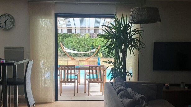 Una sala de estar con sofá, una zona de comedor con sillas y una mesa, una hamaca y vistas a una piscina y vegetación al exterior.