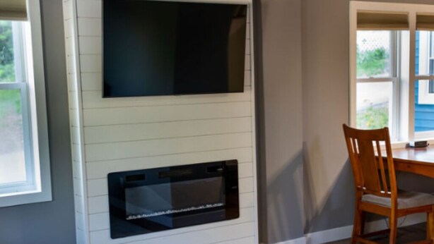 En stue med et fladskærms-tv monteret på en hvid væg, en indbygget pejs, et spiseområde med bord og stole, samt udsigt til det fri.