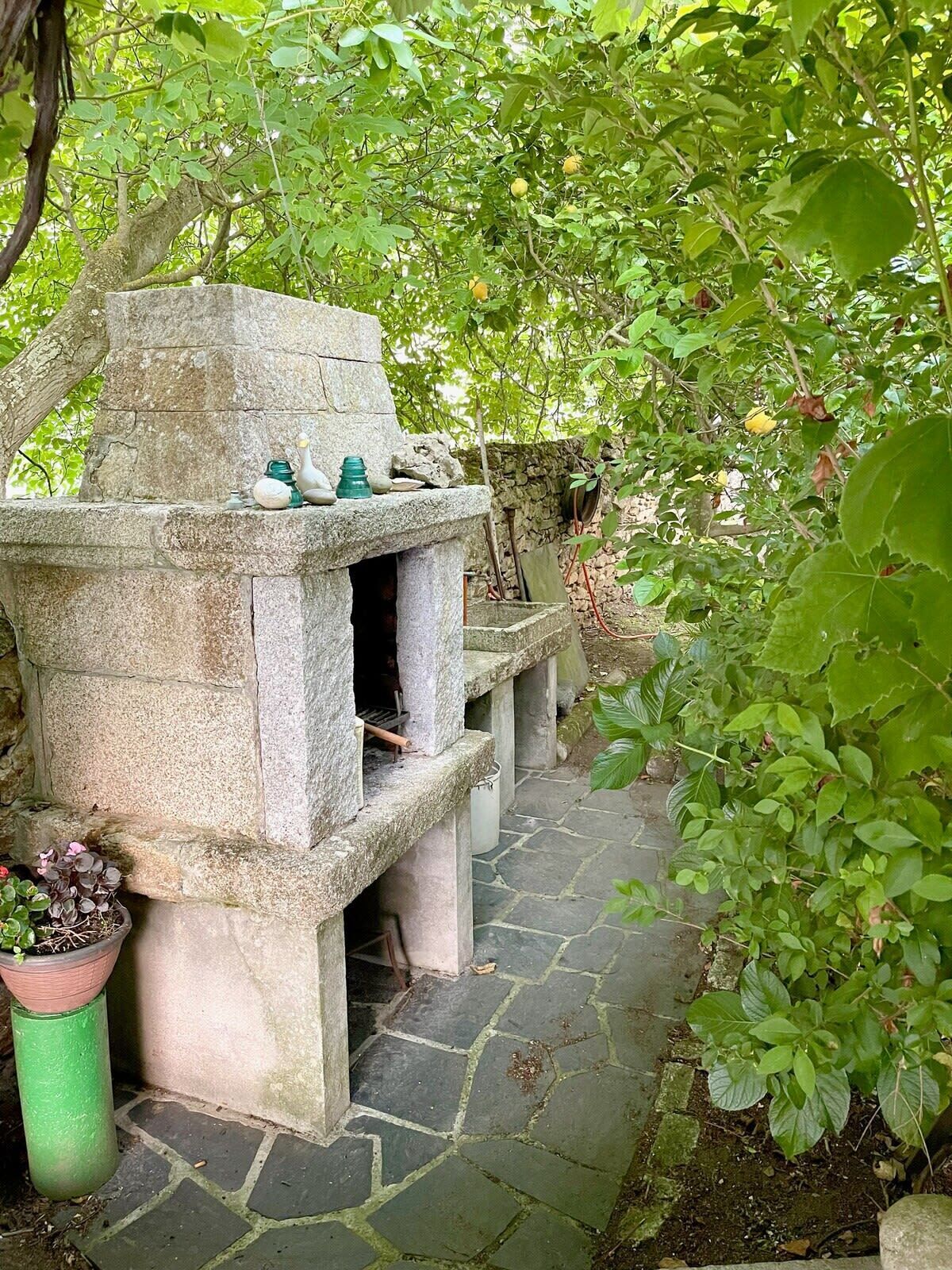 A stone-built structure with a chimney, possibly a fireplace, surrounded by lush greenery and a stone pathway.