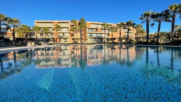 A swimming pool with clear blue water, surrounded by palm trees and modern buildings with multiple balconies. - Herdade dos Salgados, T3 8C-0C, Vila das Lagoas (Albufeira)