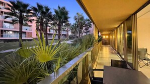 A balcony with a glass railing overlooking a landscaped area with palm trees and a building in the background. - Herdade dos Salgados, T3 8C-0C, Vila das Lagoas (Albufeira)