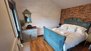 A bedroom with a wooden floor, a bed with white linens, a dresser, a mirror, and a stone wall.