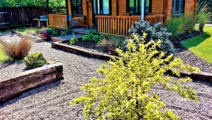 A wooden cabin with a porch, surrounded by a garden with various plants and a gravel pathway.