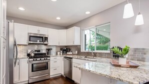 A modern kitchen with white cabinets, stainless steel appliances, a granite countertop, and a window providing a view of greenery.