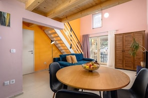 A room with a wooden staircase, a blue sofa, a round wooden table with fruit, and a view through a window.