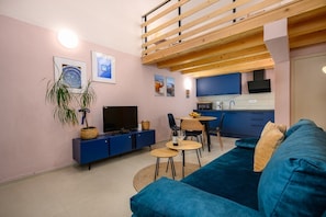 A modern living room with a blue sofa, a television, a dining table with chairs, and a kitchen area with blue cabinets.