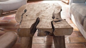 Une table en bois rustique se trouve dans un salon chaleureux, équipé d’un téléviseur, de sièges confortables et offrant une vue sur l’extérieur grâce à une grande fenêtre.