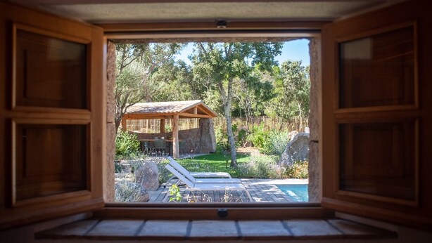 Une fenêtre à cadre en bois donnant sur un patio, qui comprend une piscine et du mobilier d’extérieur.
