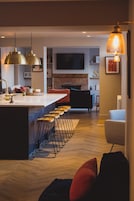 A modern kitchen with a central island, bar stools, pendant lighting, and a television in the background.