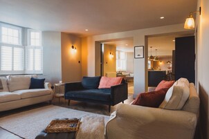 A living room with a sofa, armchairs, and a coffee table. There are windows, a door, and a kitchen area in the background.