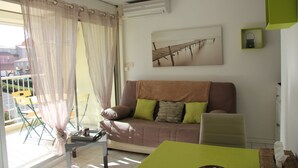 Een woonkamer met een bank, een tafel en een stoel. Er is een raam met dunne gordijnen, een schilderij aan de muur en een klein plankje met een decoratief object.