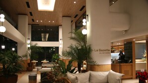 Espaço interior moderno com um sofá, plantas em vasos e uma placa com a inscrição 'La Gazette All Day Dining'.